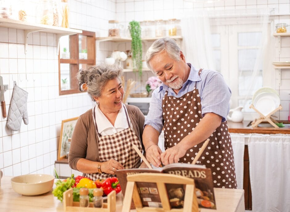 An elderly couple wearing aprons prepares a meal together in a kitchen, chopping vegetables and smiling at each other.