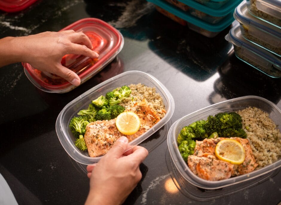 Hands arranging containers with meals of salmon, broccoli, quinoa, and lemon slices on a dark countertop.