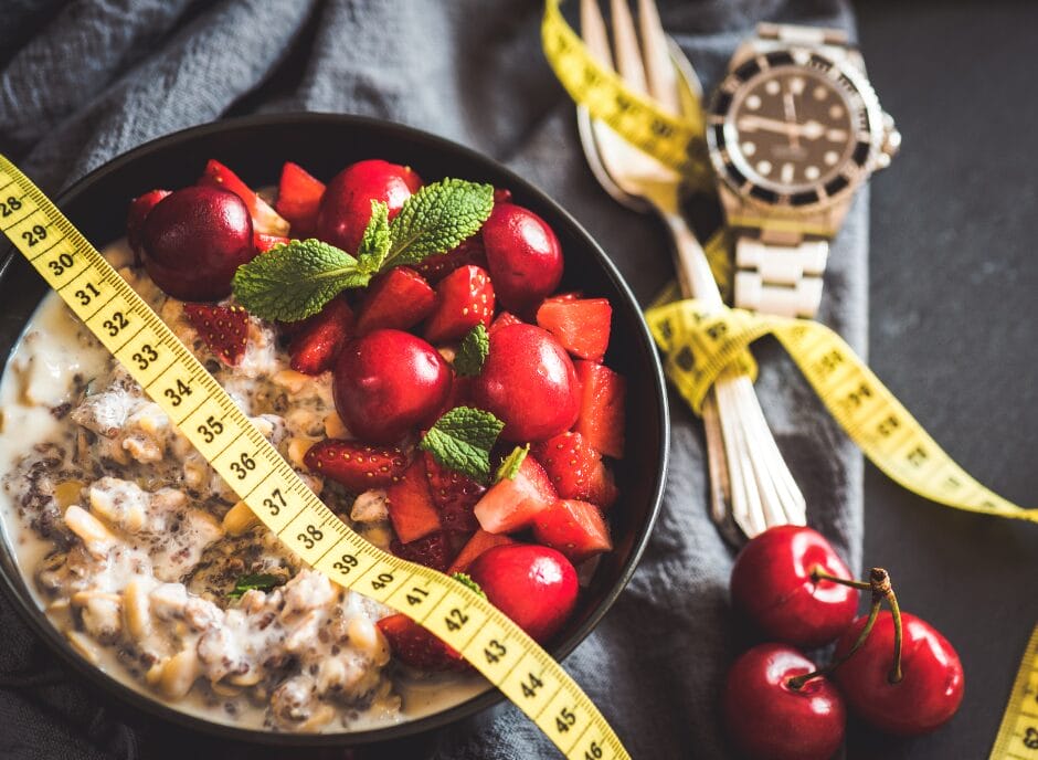 A bowl of chia pudding with strawberries and cherries, garnished with mint leaves. A tape measure wraps around, with a watch and forks nearby.