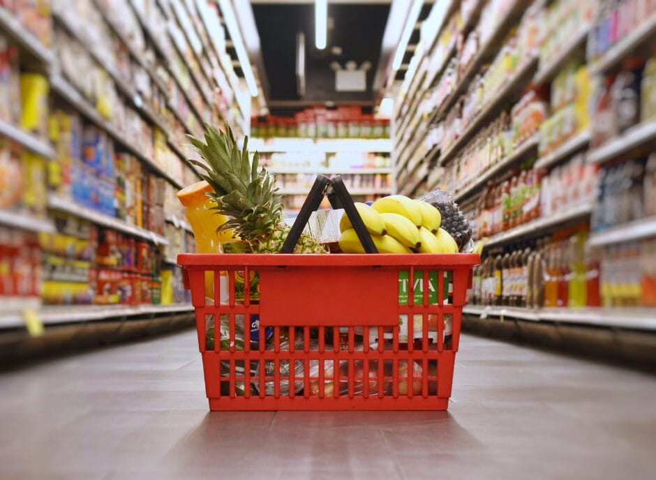 A red shopping basket filled with groceries, including bananas and a pineapple, is placed on the floor of a supermarket aisle.