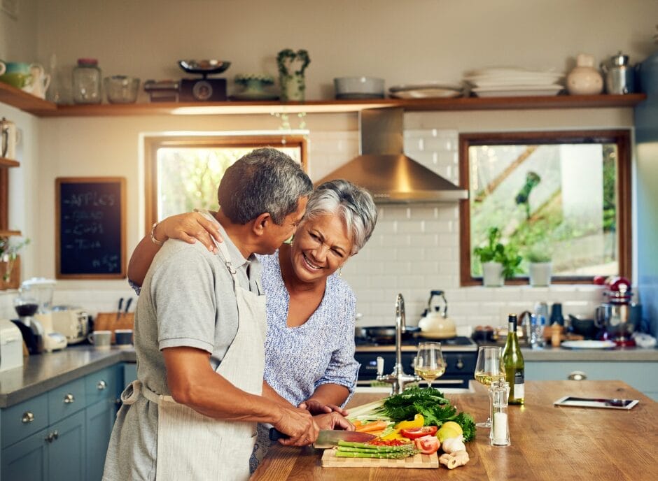 A couple is in a kitchen. The man is chopping vegetables on a wooden board while the woman smiles beside him. There are two wine glasses and bottles on the counter.