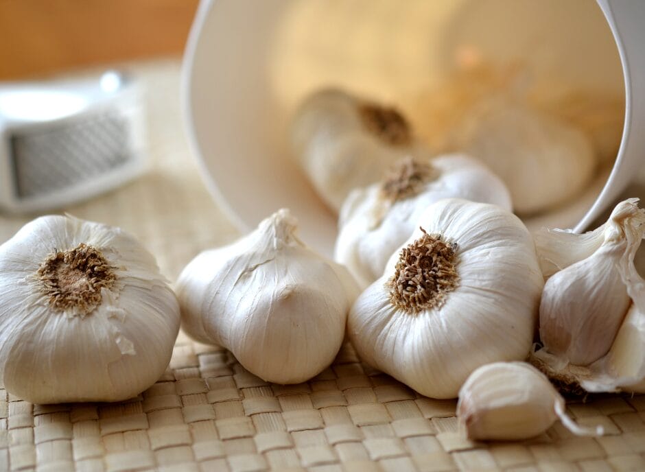 Whole garlic bulbs and cloves spill from a white container onto a woven surface.