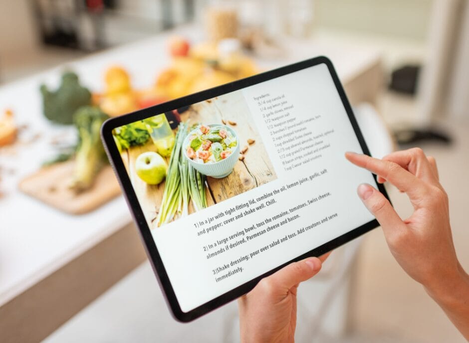 Person holding a tablet displaying a salad recipe, with a kitchen counter and various vegetables in the background.