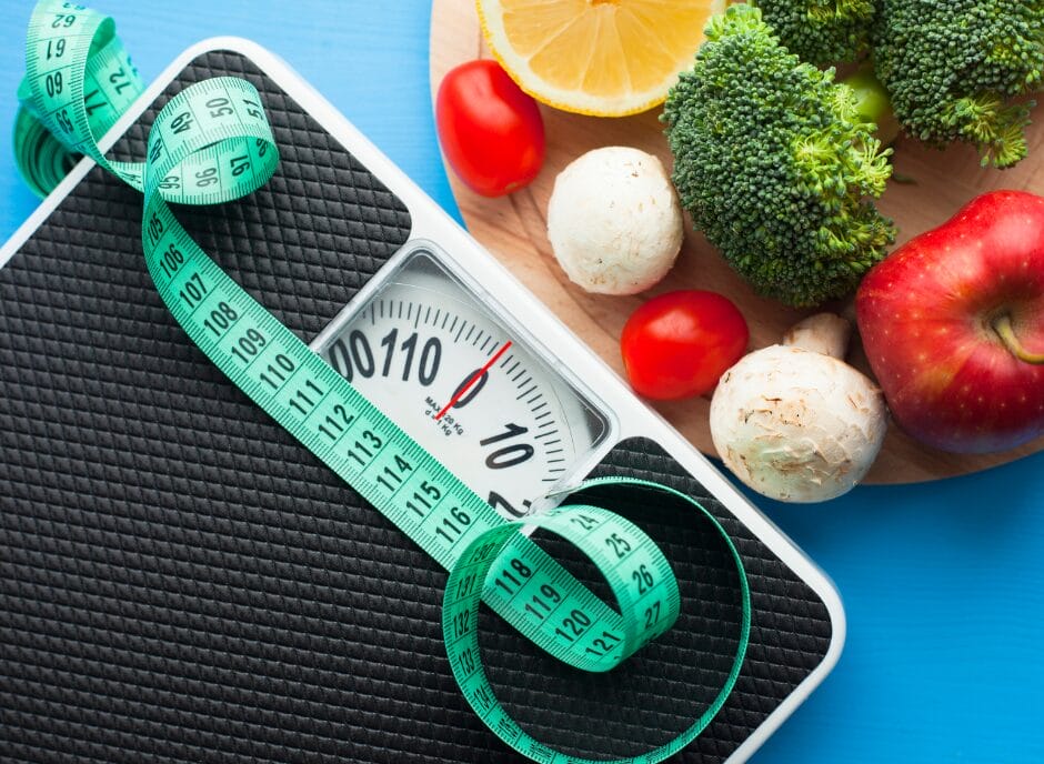 A weighing scale with a measuring tape on it is next to a plate of vegetables and fruits, including broccoli, tomatoes, mushrooms, lemon, and an apple, on a blue background.