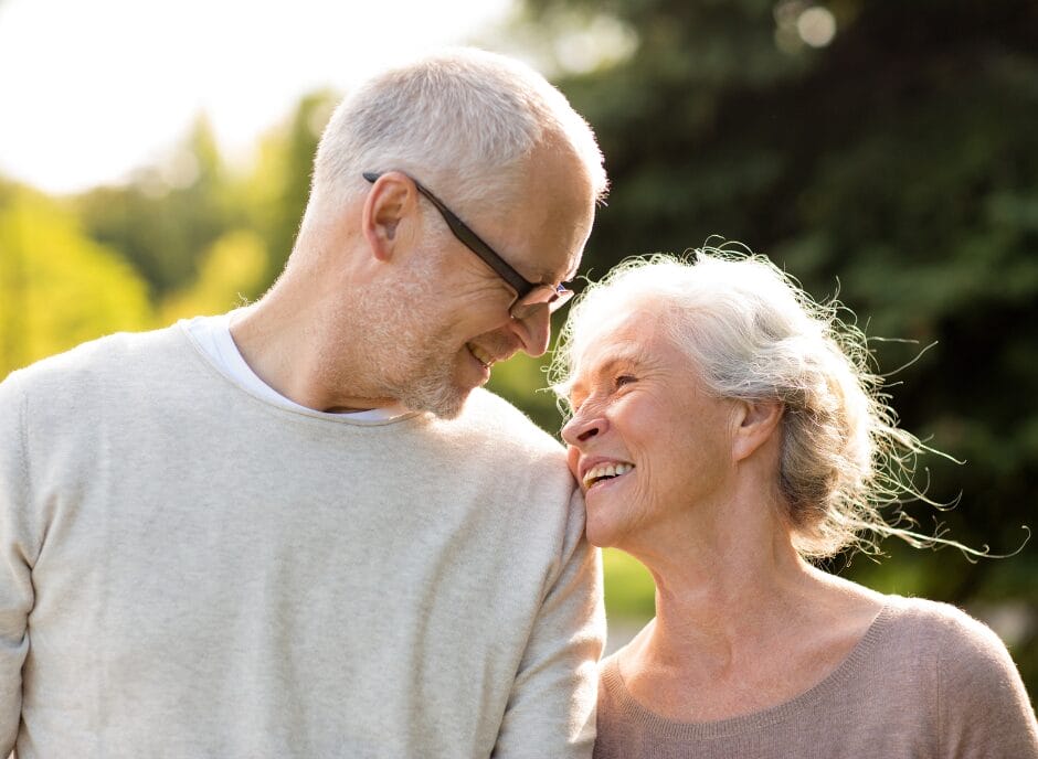 An older couple smiling at each other outdoors, with greenery in the background.