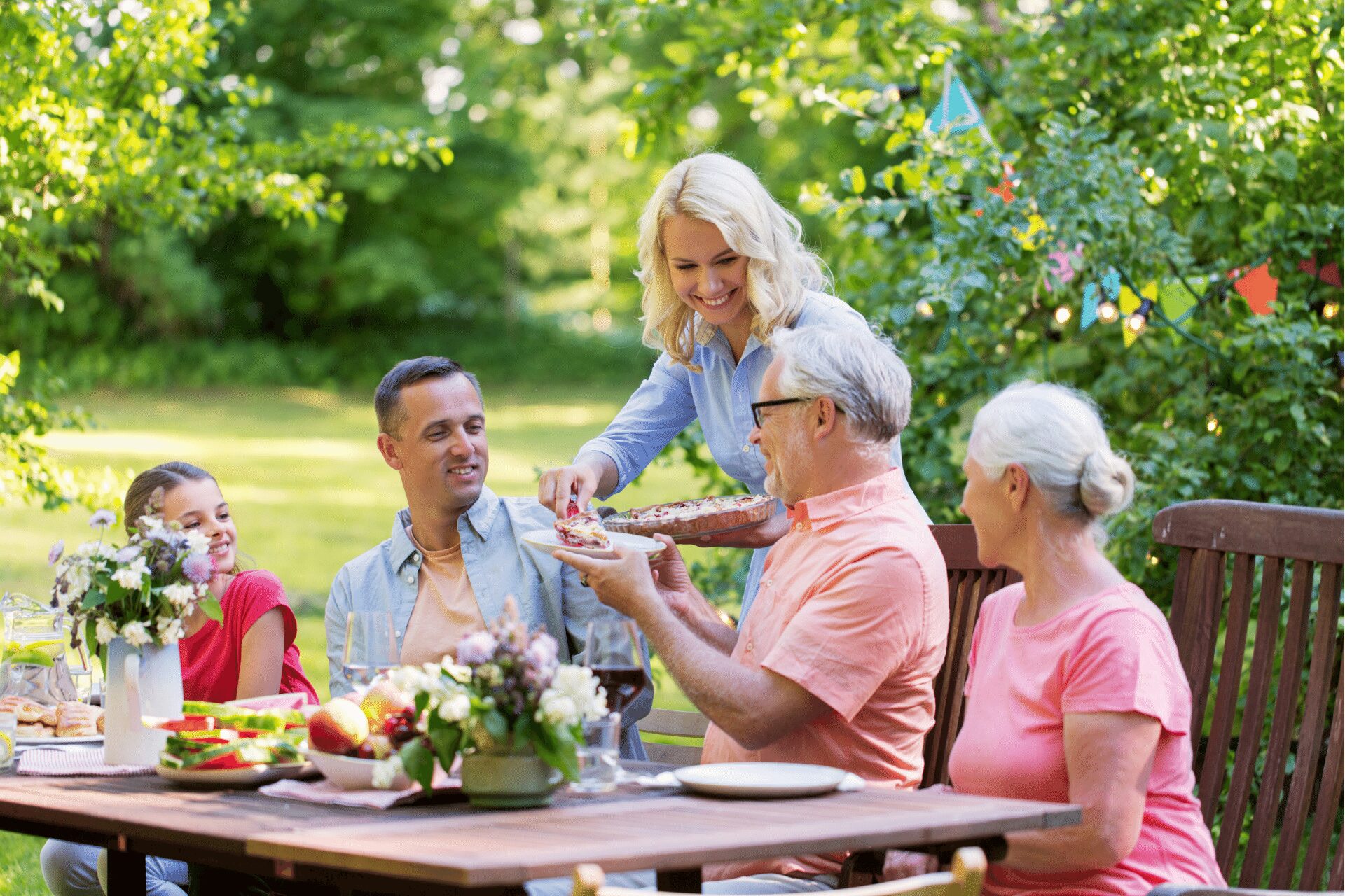 A group of people dining outdoors at a table in a garden setting. One person is serving food to others. The table is set with flowers, fruit, and drinks. Trees and decorations are in the background. A group of people dining outdoors at a table in a garden setting. One person is serving food to others. The table is set with flowers, fruit, and drinks. Trees and decorations are in the background.