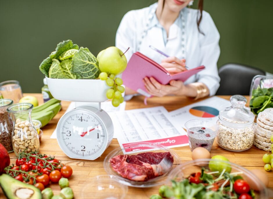 A nutritionist specializing in how to find a renal dietitian takes notes with various healthy foods, including fruits, vegetables, and meat, spread on a table, with a focus on a balanced diet.