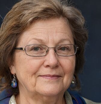 A woman with short, grayish-brown hair and glasses, wearing a beige top and blue earrings, is looking at the camera against a dark background.