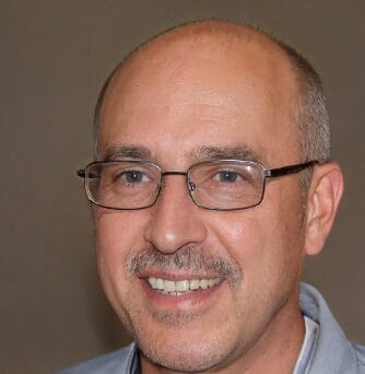 A man with glasses, a mustache, and short-cropped hair smiles at the camera. He is wearing a light blue shirt.