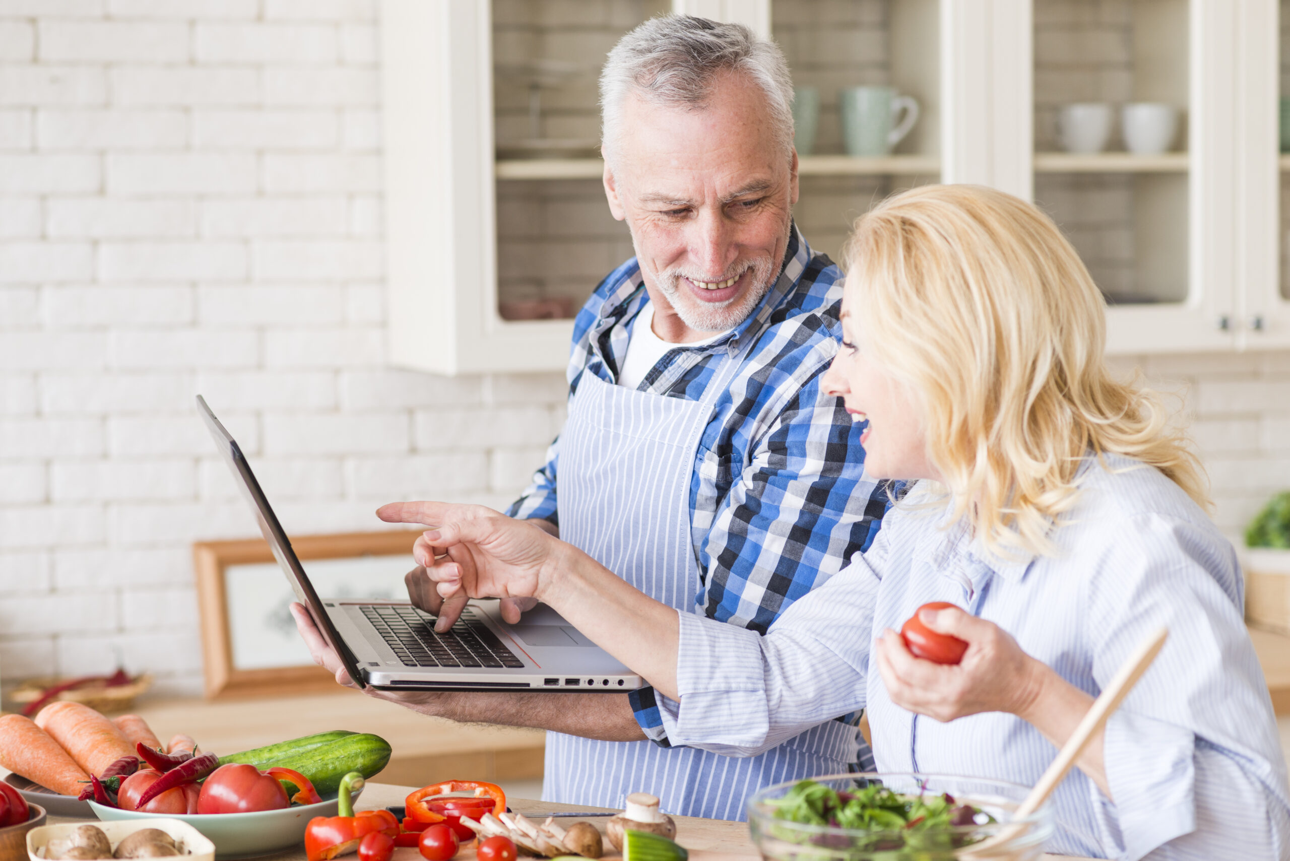 smiling-senior-man-looking-her-wife-pointing-laptop-while-preparing-salad-kitchen