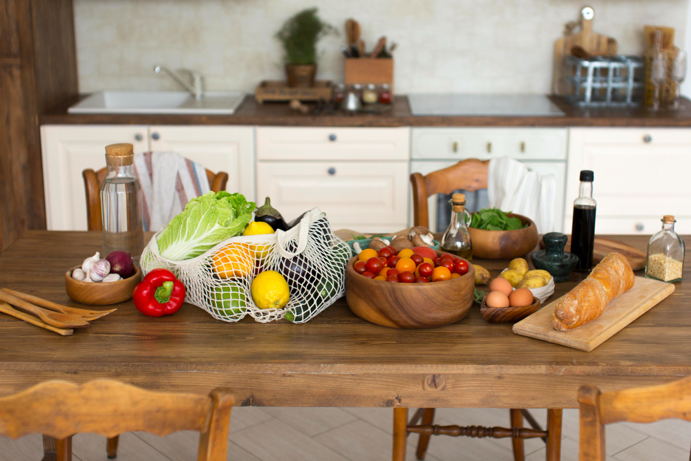 A wooden kitchen table with various fresh vegetables, eggs, a baguette, bowls, bottles, and a net shopping bag, set in a rustic kitchen. A wooden kitchen table with various fresh vegetables, eggs, a baguette, bowls, bottles, and a net shopping bag, set in a rustic kitchen.
