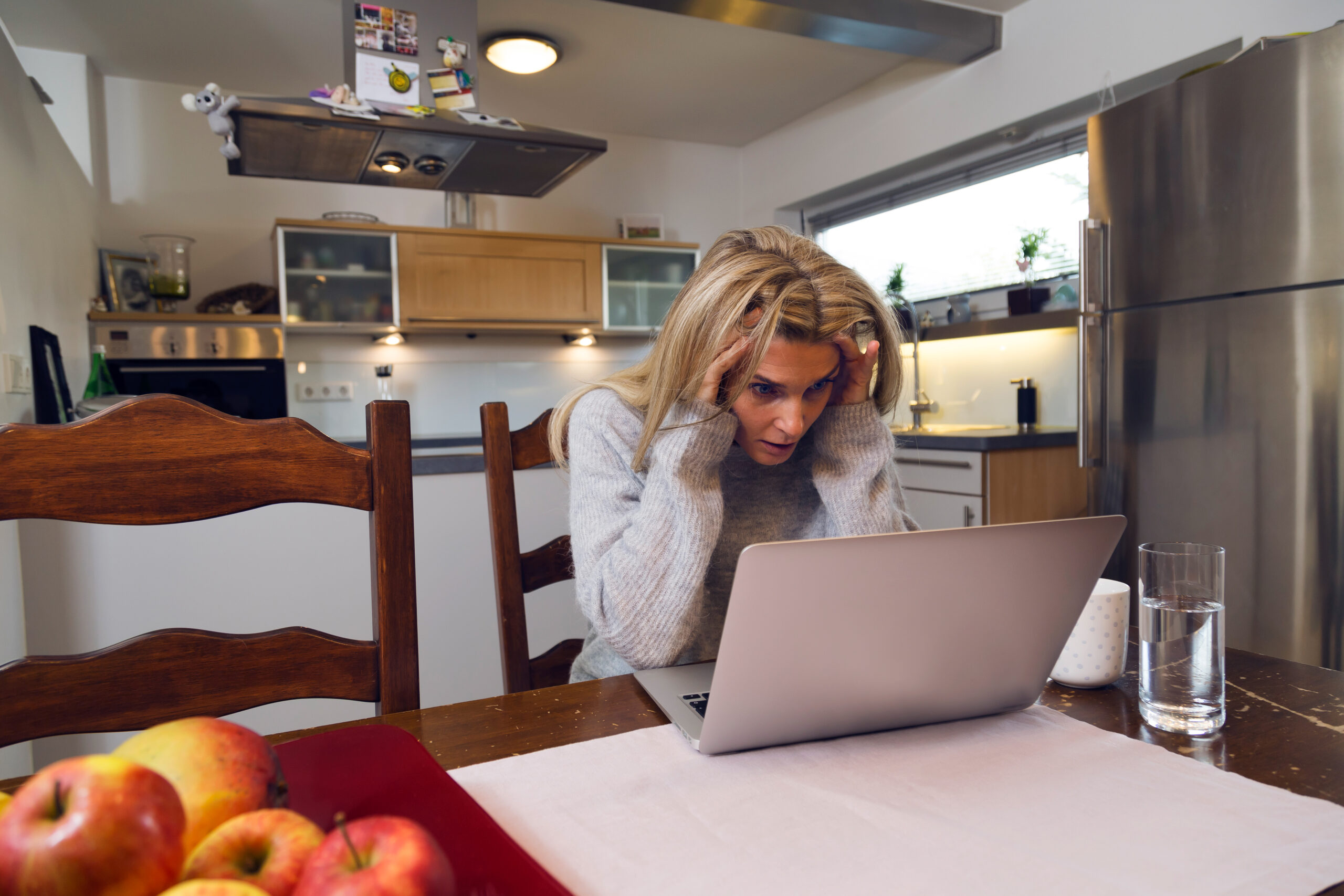 A woman sits at a kitchen table looking stressed while using a laptop, with a cup, glass of water, and fruit bowl on the table. A woman sits at a kitchen table looking stressed while using a laptop, with a cup, glass of water, and fruit bowl on the table.