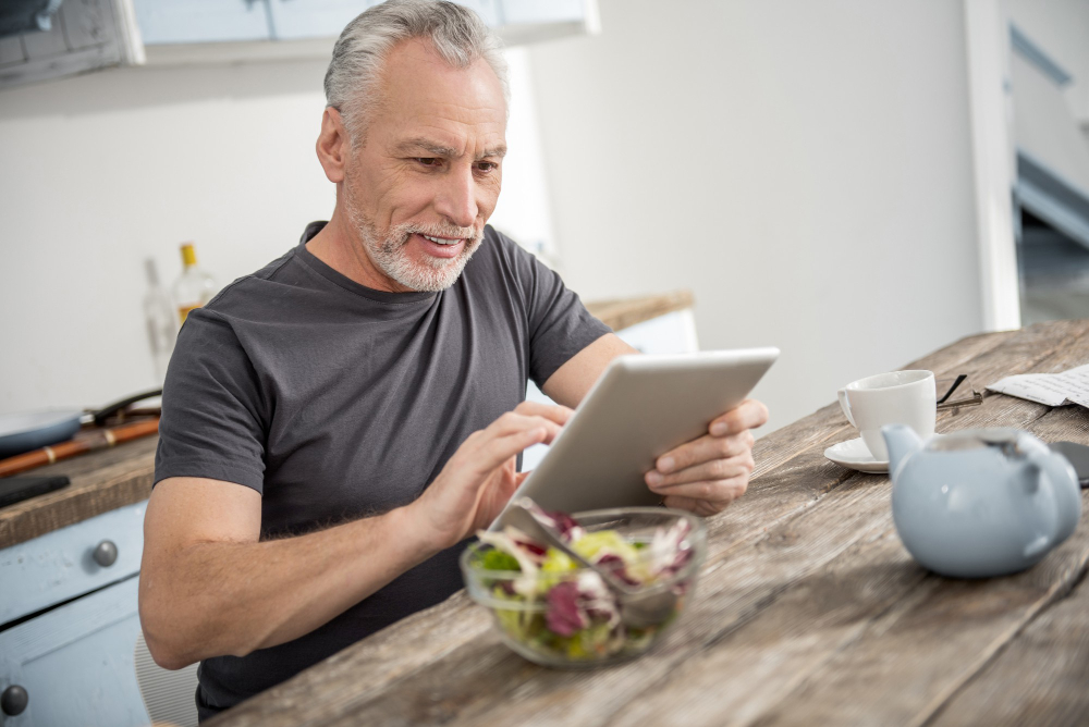 A man sits at a wooden kitchen table using a tablet, with a bowl of salad, a teapot, and a cup nearby. A man sits at a wooden kitchen table using a tablet, with a bowl of salad, a teapot, and a cup nearby.