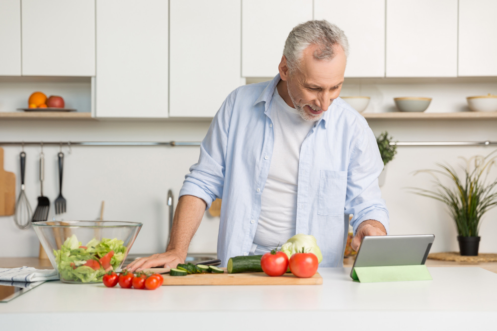 A man in a light blue shirt stands in a modern kitchen, looking at a tablet while preparing vegetables on a cutting board. A man in a light blue shirt stands in a modern kitchen, looking at a tablet while preparing vegetables on a cutting board.