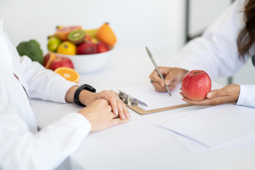 Two people in white coats sit at a table with a clipboard; one holds an apple while the other writes. A bowl of assorted fruit is in the background. Two people in white coats sit at a table with a clipboard; one holds an apple while the other writes. A bowl of assorted fruit is in the background.