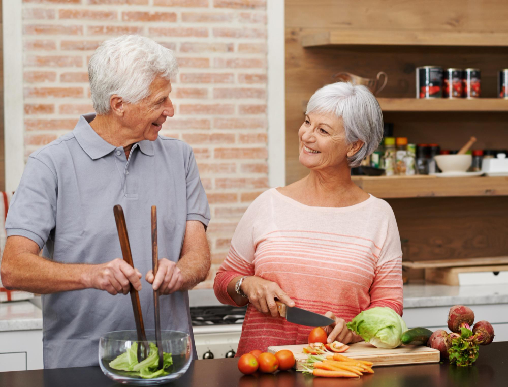 Two older adults stand in a kitchen preparing a salad together, smiling at each other. Vegetables and utensils are on the counter in front of them.