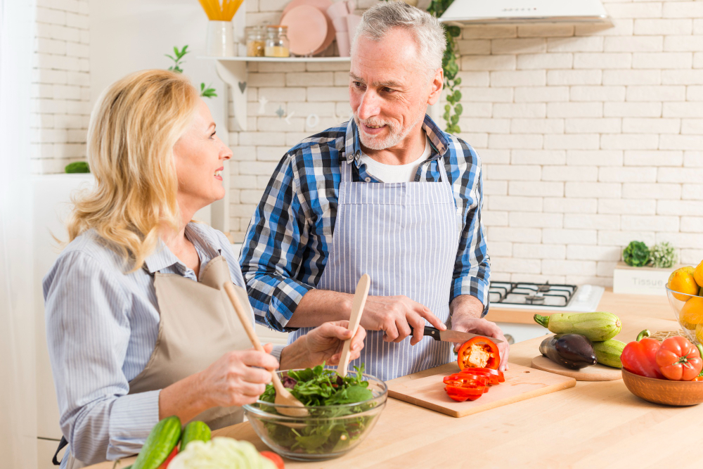 An older man chops tomatoes while a woman mixes salad greens; both wear aprons and smile at each other in a bright kitchen with fresh vegetables on the counter.