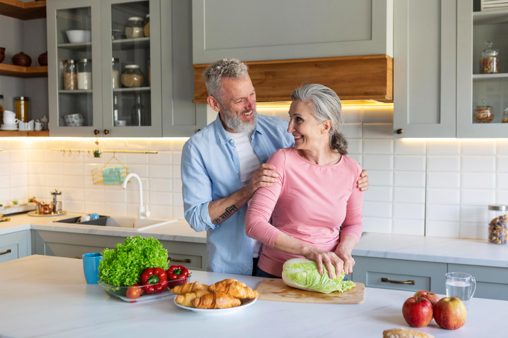 An older couple stands in a kitchen; the woman chops cabbage while the man stands behind her, smiling. Fresh vegetables, croissants, and apples are on the counter.