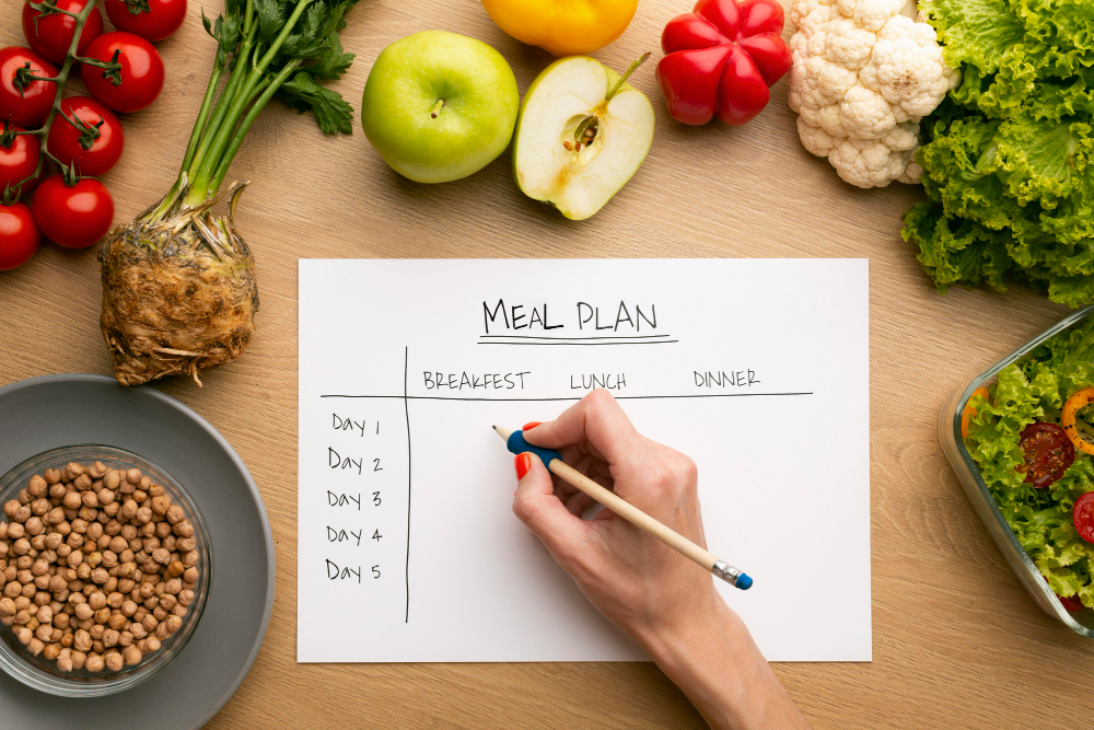 A hand writes on a meal plan chart surrounded by various fresh vegetables, fruit, chickpeas, and a bowl of salad on a wooden table.