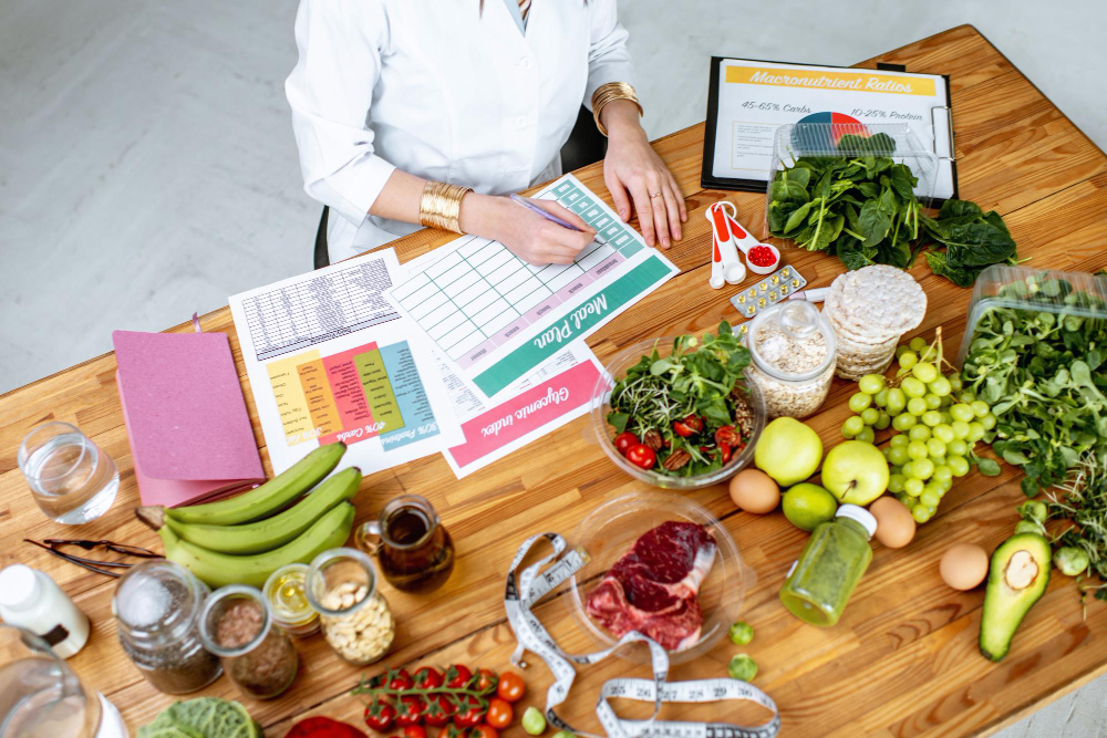 A person at a table reviews nutrition charts surrounded by healthy foods like fruits, vegetables, nuts, meat, and measuring tools.