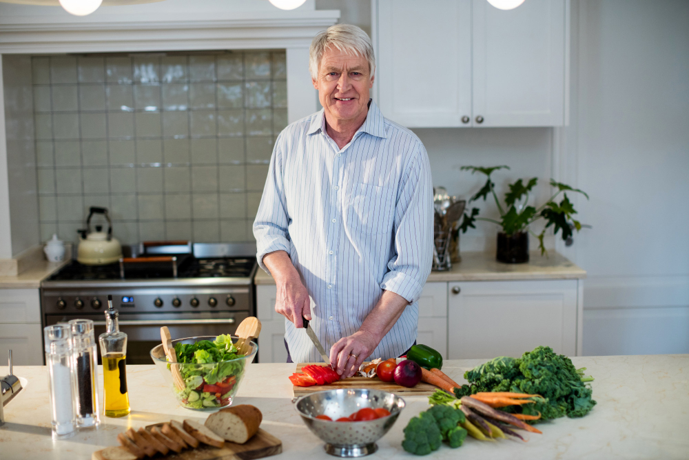 An older man is chopping vegetables on a kitchen counter with various fresh produce, bread, and salad ingredients around him.