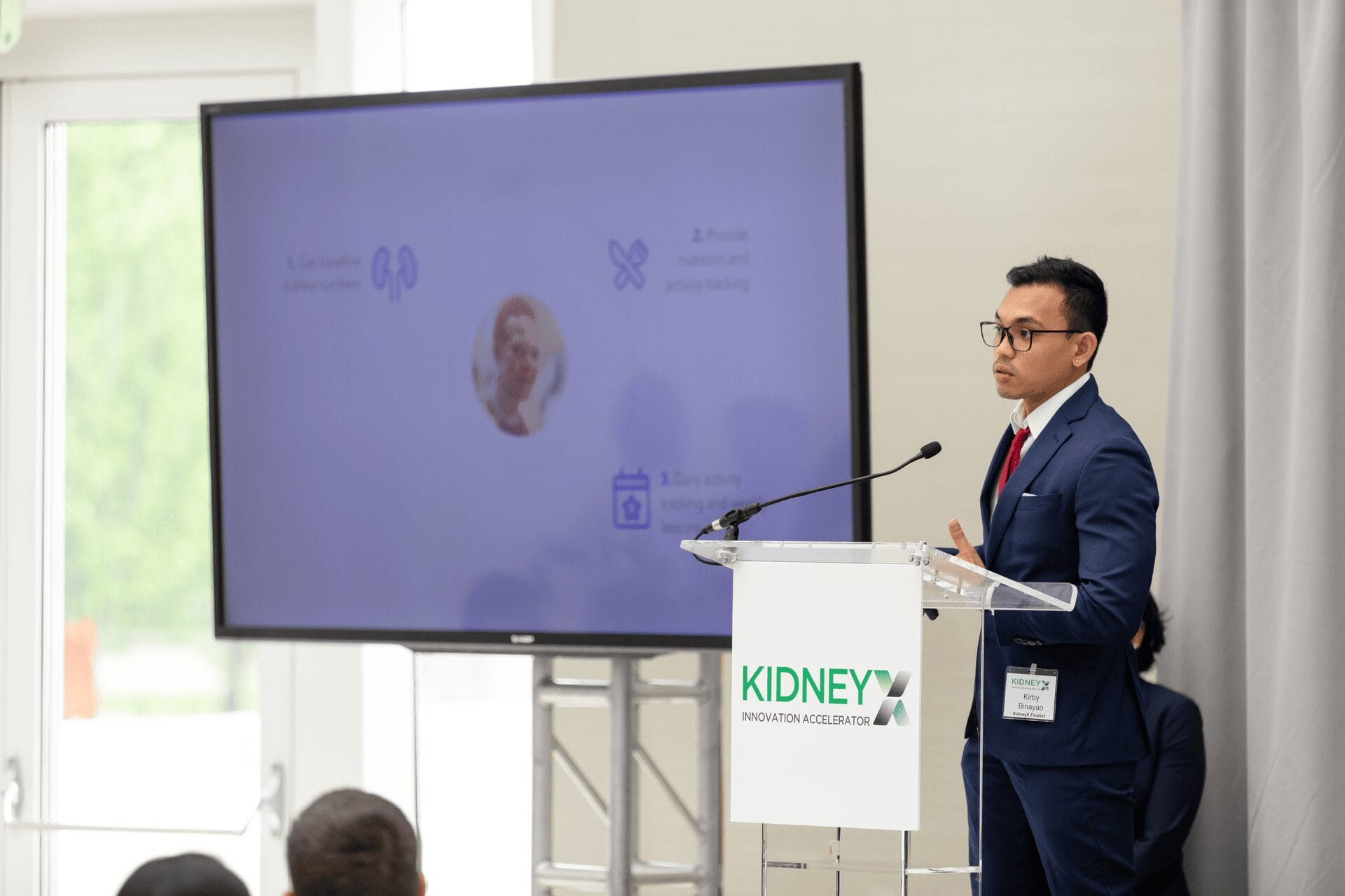 A man in a suit stands at a podium labeled "Kidney Innovation Accelerator," presenting a slide to an audience.