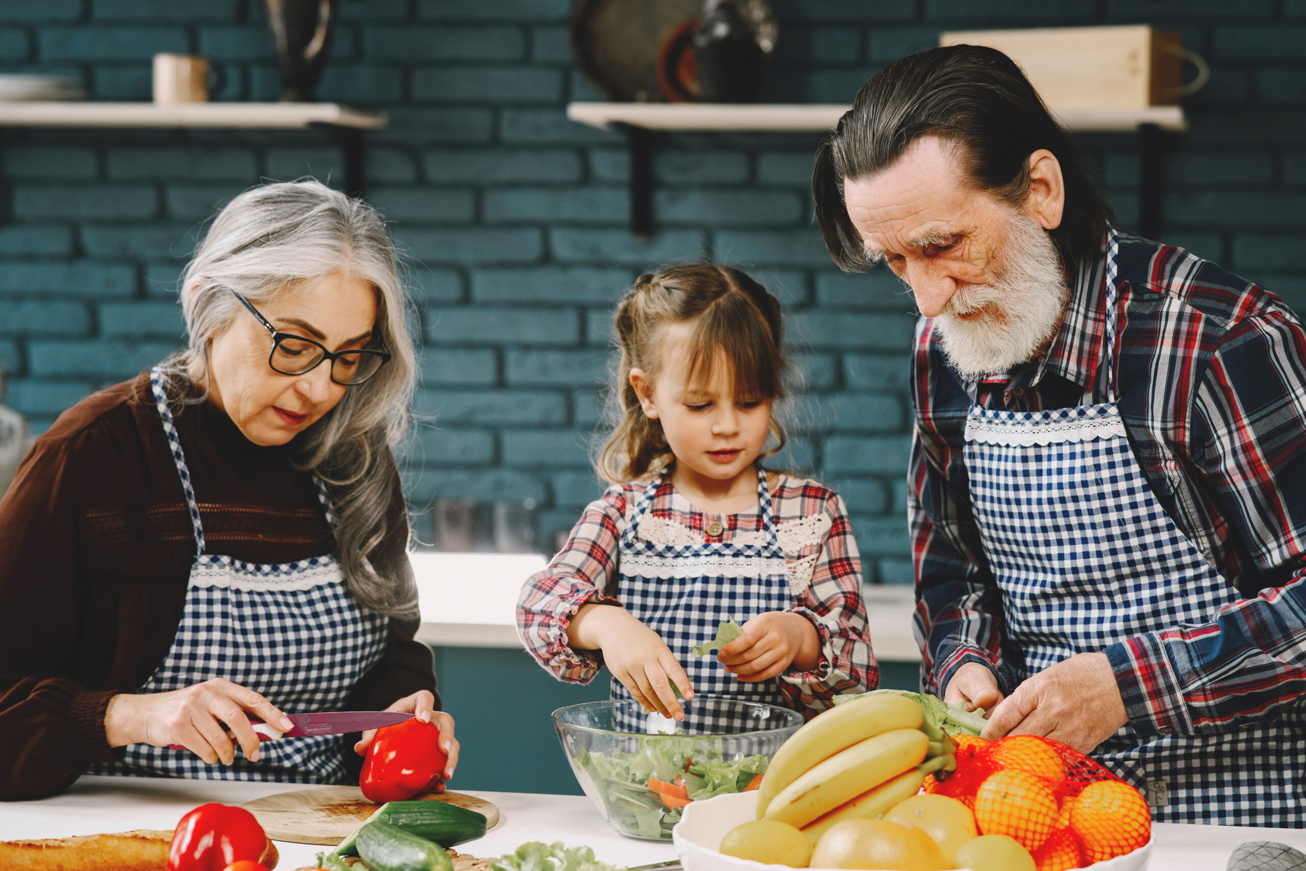 An older woman, a young girl, and an older man wearing aprons prepare fruits and vegetables together in a kitchen.