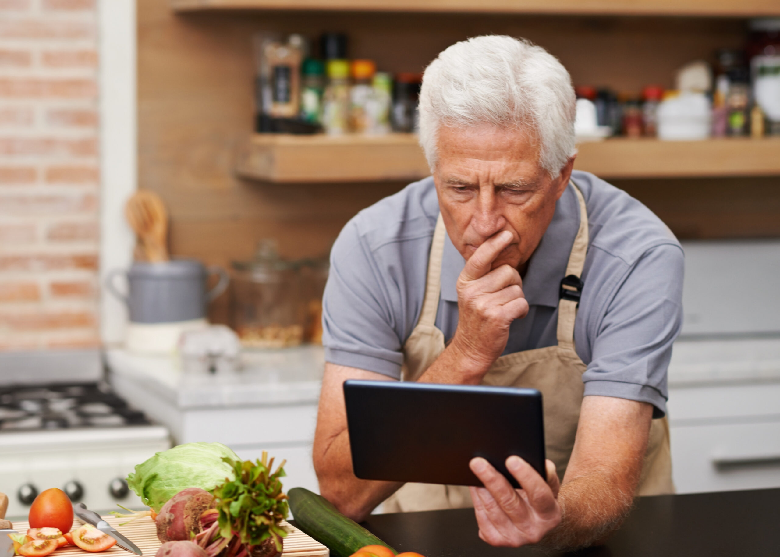 An older man wearing an apron looks at a tablet in a kitchen with vegetables on the counter.