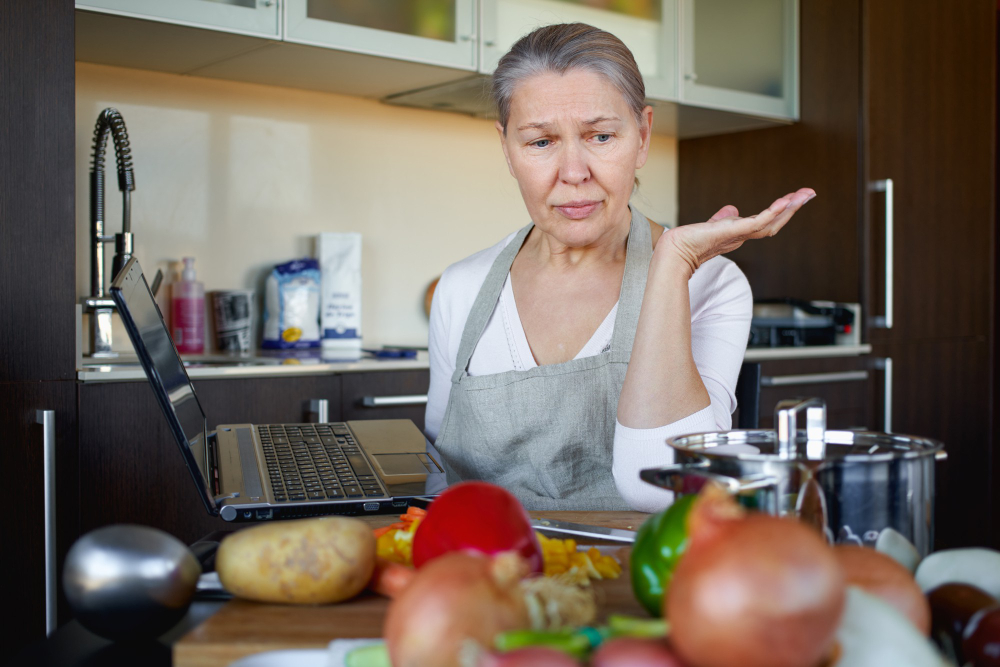 Woman in apron looking confused at a laptop in a kitchen, surrounded by various vegetables and cooking utensils on the counter.