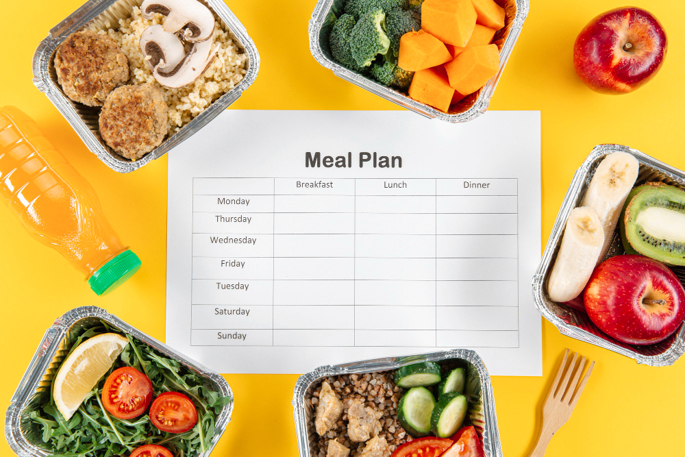 A blank meal plan template is surrounded by healthy foods in containers, a bottle of juice, an apple, and various fruits and vegetables on a yellow background.