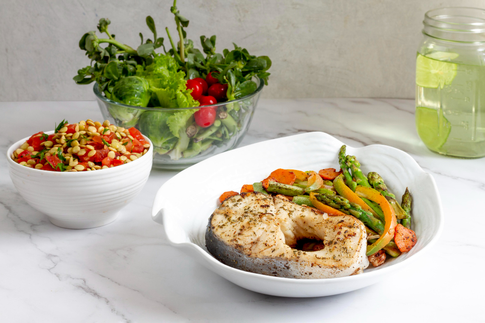 A plate with a cooked fish steak, sautéed vegetables, a bowl of salad with pine nuts, a glass of water with lime, and a bowl of mixed greens on a white surface.
