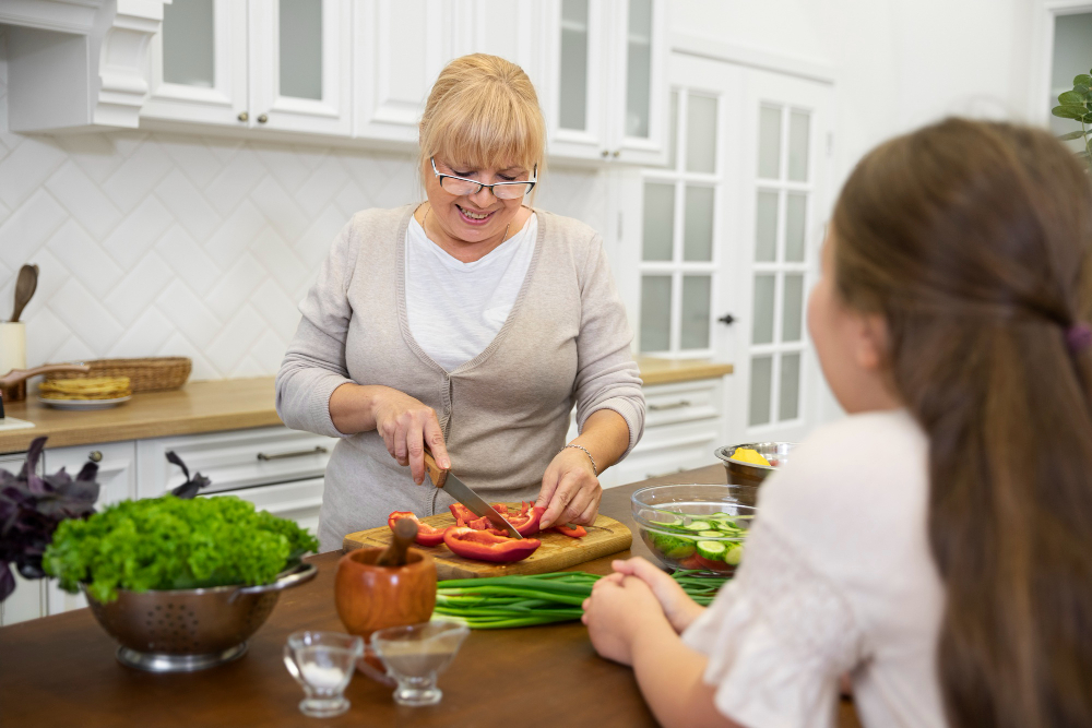 An older woman slices red bell pepper on a cutting board while a young girl watches at a kitchen counter with fresh vegetables.