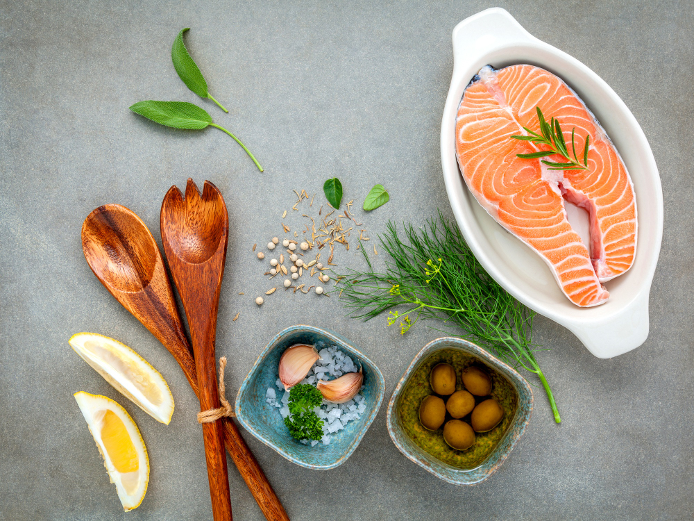Raw salmon steak in a white dish with lemon wedges, wooden utensils, garlic, herbs, olives, and seasoning arranged on a gray surface.