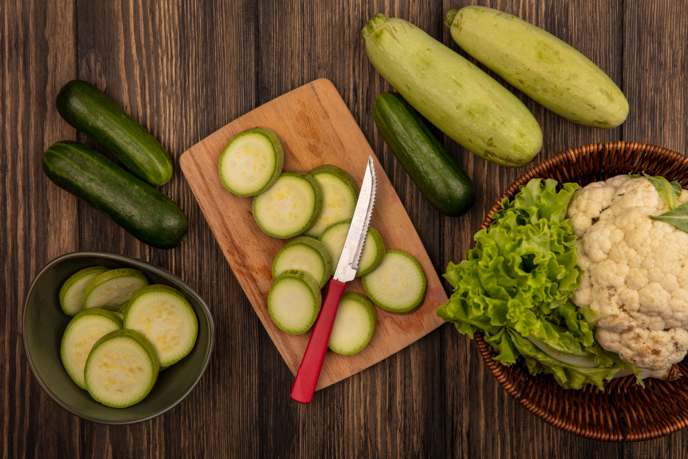 A cutting board with sliced zucchini and a red-handled knife, surrounded by whole zucchinis, lettuce, and cauliflower on a wooden table.